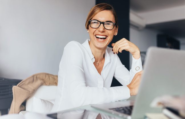 Business woman in spectacles sitting at her desk in office and laughing. Woman sitting in office with laptop computer on her desk.